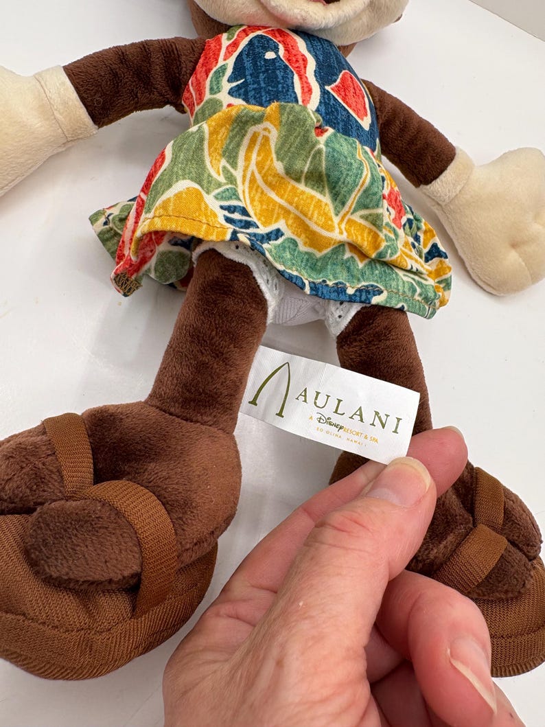 May include: A plush doll wearing a colorful floral print dress and brown sandals. The doll has brown arms and legs, with white gloves and a white face. A tag reads "Aulani, A Disney Resort & Spa".