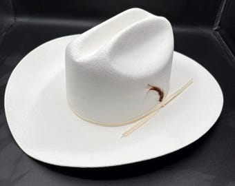 Puede incluir: Sombrero de vaquero blanco con ala ancha y copa alta. El sombrero presenta una banda de color claro y un pequeño adorno de pluma. Fabricado con material tejido, adecuado para la protección solar.