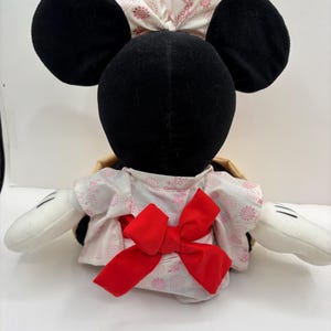 May include: A plush toy featuring a black head with large round ears and white gloves. The toy wears a white and pink floral patterned kimono with a large red bow. A matching bow adorns the head.
