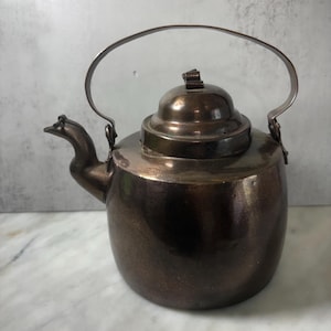 May include: A dark metallic tea kettle with a rounded body, a curved spout, and a hinged handle. The kettle features a removable lid with a small knob on top. The surface has a polished, reflective finish with visible signs of aging and patina.