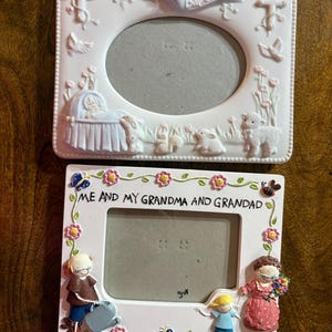 以下が含まれることがあります： 2つの白いフォトフレーム。上部のフレームには「Baby's Baptism」と書かれたバナーがあり、揺りかごの中の赤ちゃん、子羊、ウサギが描かれています。下のフレームには「ME AND MY GRANDMA AND GRANDAD」と書かれており、家族の姿が描かれています。