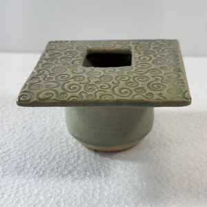 May include: A handcrafted ceramic vase with a square top and a circular base. The top features a square opening and is decorated with a swirling pattern. The vase is a muted green color, with a natural, earthy aesthetic.