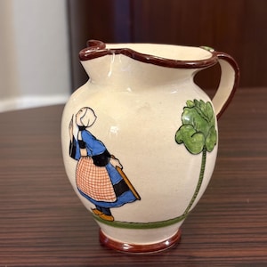 May include: A small, cream-colored ceramic pitcher with a brown rim and handle. The pitcher features a hand-painted illustration of a person in traditional clothing and a green plant. The base and rim are brown.