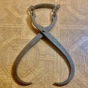 Puede incluir: Tenazas de hielo de metal antiguas con un asa curvada y puntas. Las tenazas son de color gris oscuro y tienen un diseño cruzado. El asa está conectada a las tenazas con cadenas de metal.