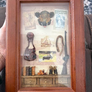 May include: A framed shadow box with a hunting theme. It features miniature figurines of a boar's head, a rifle, a hunting bag, binoculars, books, and a hunting scene. The frame is made of dark brown wood.