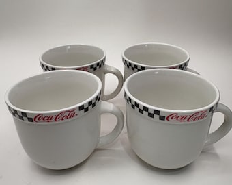Op de afbeelding: Vier witte keramische Coca-Cola-mokken. Elke mok heeft een zwart-wit geblokte band rond de rand en het Coca-Cola-logo in rood. De mokken hebben een afgeronde vorm met een oor.