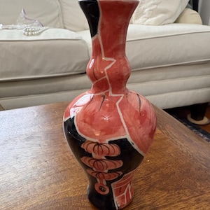 Op de afbeelding: Een keramische vaas met een bolvormig lichaam en een smalle hals, voorzien van een handgeschilderd ontwerp in koraalroze en zwart. Het oppervlak toont abstracte geometrische vormen en gestileerde bloemmotieven omlijnd met dunne zwarte lijnen.