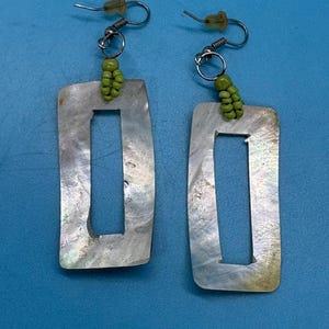 K&ouml;nnte beinhalten: Ein Paar rechteckige Ohrringe mit Perlmutt-Finish und einem mittigen Ausschnitt. Jeder Ohrring ist mit einem silberfarbenen Haken mit einer Ansammlung kleiner, gr&uuml;ner Perlen verbunden. Die Ohrringe sind vor blauem Hintergrund platziert.