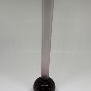 May include: A tall, slender glass vase with a bulbous base. The vase has a clear, cylindrical neck that tapers slightly towards the top. The base is a dark, purplish-brown color with a textured, bubble-like appearance. The vase is designed for a single stem flower.