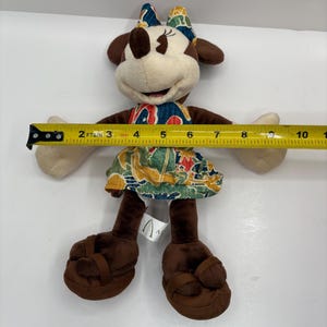 May include: A Minnie Mouse plush doll wearing a colorful dress and bow. The doll is approximately 8 inches tall, with brown ears, shoes, and arms. The dress features a tropical print with red, blue, and green accents. The doll has a smiling expression.