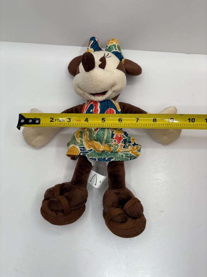 May include: A Minnie Mouse plush doll wearing a colorful dress and bow. The doll is approximately 8 inches tall, with brown ears, shoes, and arms. The dress features a tropical print with red, blue, and green accents. The doll has a smiling expression.