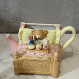 Könnte beinhalten: Eine skurrile Keramik-Teekanne in Form eines Bettes, mit einem Teddybär-Kopf und kleinem Zubehör. Die Teekanne ist hauptsächlich beige, rosa und gelb, mit einer floralen Bettdecke. Der Griff und der Ausguss sind gebogen.