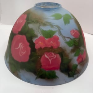 Può includere: Un paralume in vetro con un motivo floreale dipinto. Il paralume presenta rose rosa e foglie verdi su uno sfondo azzurro. Il paralume ha una forma arrotondata e un'apertura leggermente svasata.