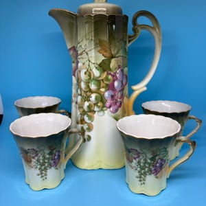 May include: A vintage porcelain coffee set featuring a tall coffee pot with a lid and four matching cups. The set is painted with a grape vine design in shades of green, purple, and yellow. The cups have gold trim and handles.
