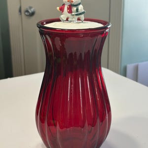 Op de afbeelding: Een robijnrode glazen vaas met verticale geribbelde details. De vaas is bekroond met een wit oppervlak en twee sneeuwpopfiguren. De sneeuwpoppen dragen winterkleding, waaronder hoeden en sjaals. De vaas is waarschijnlijk voor decoratieve doeleinden.