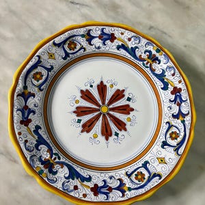 Op de afbeelding: Een decoratieve keramische plaat met een geschulpte gele rand. De plaat heeft een wit midden met een bloemmotief in rood, bruin en geel. De rand heeft blauwe en gele bloempatronen.