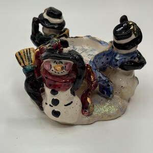 May include: A ceramic candle holder featuring three snowman figures. The snowmen wear black hats and scarves, encircling a central bowl. The snowmen are painted with coal eyes and button details. The base has a glittery finish, adding a festive touch.