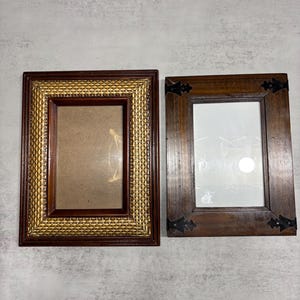 May include: Two rectangular picture frames. One frame has a woven gold and brown border, and the other is a dark brown wood frame with black corner accents. Both frames have a rectangular opening for a photo.