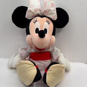 May include: A plush Minnie Mouse doll with black ears, a pink face, and a large bow with a floral pattern. She wears a white dress with a pink floral design and a red sash. Her large feet are beige with black soles.