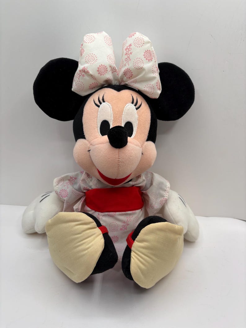 May include: A plush Minnie Mouse doll with black ears, a pink face, and a large bow with a floral pattern. She wears a white dress with a pink floral design and a red sash. Her large feet are beige with black soles.