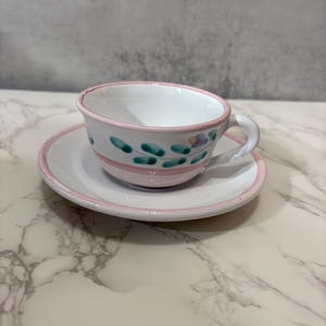May include: A white ceramic teacup and saucer set. The cup and saucer have a pink rim and are decorated with green and blue floral designs. The cup has a curved handle.