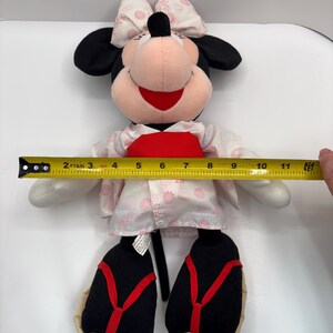 May include: A Minnie Mouse plush doll wearing a kimono. The doll has black ears, a pink face, and a red mouth. The kimono is white with pink floral patterns and a red obi. The doll measures approximately 10 inches in height.