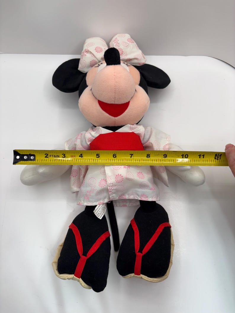 May include: A Minnie Mouse plush doll wearing a kimono. The doll has black ears, a pink face, and a red mouth. The kimono is white with pink floral patterns and a red obi. The doll measures approximately 10 inches in height.