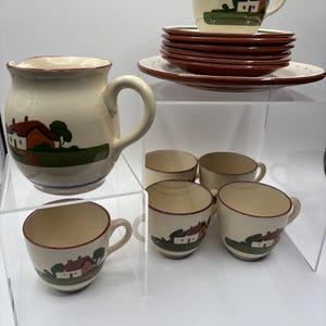 May include: A cream-colored ceramic tea set featuring a house motif. The set includes a pitcher, cups, saucers, and teacups. The house design features a red roof, green grass, and a blue stripe. The rims of the plates and cups are brown.