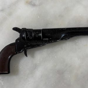 May include: A miniature toy revolver with a black metal barrel and frame featuring the embossed text OLD TIGER. The handle is a dark brown colour with a smooth finish. The toy gun has a hammer, trigger, and cylinder visible on the side.