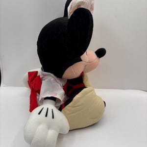 May include: A plush Minnie Mouse doll, featuring black ears, a pink nose, and a cream-colored face. She wears a kimono-style outfit with a red bow and a patterned headpiece. The doll's white gloves and tan shoes are also visible.