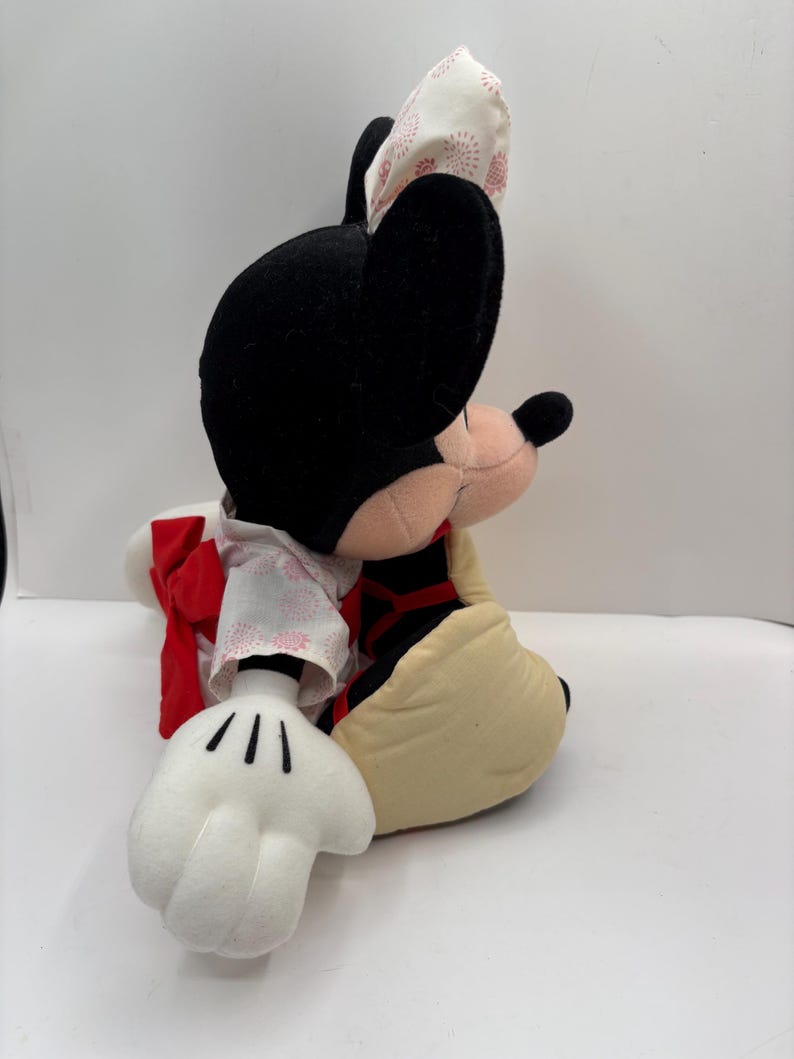 May include: A plush Minnie Mouse doll, featuring black ears, a pink nose, and a cream-colored face. She wears a kimono-style outfit with a red bow and a patterned headpiece. The doll's white gloves and tan shoes are also visible.