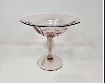 May include: A vintage pink glass pedestal bowl with a wide, shallow rim and a rounded stem. The bowl has a circular base and is likely used for serving desserts or displaying decorative items. The glass has a slight iridescence.