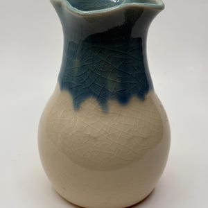 May include: A ceramic vase with a bulbous base and a flared, ruffled rim. The vase features a two-tone color scheme, with a cream-colored base and a dark blue, crackled glaze that transitions to a lighter blue at the rim. The vase is approximately 6 inches tall.