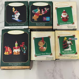 Pode incluir: Uma coleção de miniaturas de Natal em caixas. Os enfeites apresentam personagens como Snoopy, Ursinho Pooh e Papai Noel. As caixas são verdes e douradas, com o texto "Keepsake Ornament" e "Miniature Collector's Series."