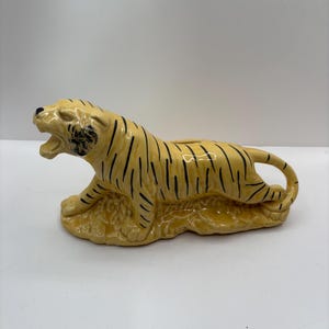 May include: A ceramic tiger figurine in a reclining pose, painted in a light yellow with black stripes. The tiger has an open mouth and is resting on a textured base. The figurine is a decorative item.