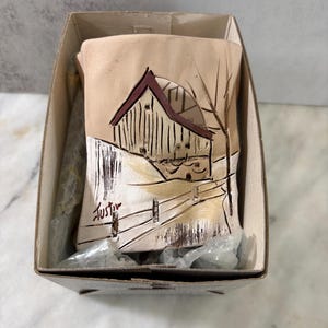 May include: A rectangular ceramic plaque featuring a hand-painted scene of a barn in a snowy landscape with a fence and bare trees. The artwork is rendered in shades of tan, white, and dark brown, with the signature Justin visible in the lower left corner.