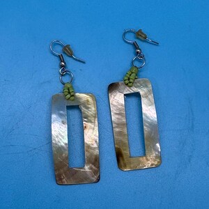 May include: A pair of rectangular earrings with a mother-of-pearl finish. The earrings have a cutout rectangle in the center and are attached to silver-colored hooks with small green beads. The background is a solid blue.