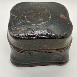 Puede incluir: Una caja cuadrada de cerámica marrón oscuro con tapa. La caja tiene un acabado brillante y una forma ligeramente irregular. La tapa presenta acentos moteados de color marrón rojizo y crema. La caja parece hecha a mano.