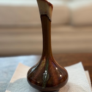 Könnte beinhalten: Eine hohe, schlanke Keramikvase mit bauchigem Fuß und einem schmalen, langgestreckten Hals. Das Stück hat eine glänzende, dunkelbraune Glasur mit helleren, cremefarbenen Tropfen am Rand und dezenten grünen vertikalen Streifen am Körper.