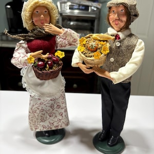 Pode incluir: Duas figuras decorativas de um homem e uma mulher. A mulher usa um chapéu branco, um corpete vermelho e um vestido com estampa floral, segurando uma cesta de frutas vermelhas. O homem usa um boné e um colete de tweed, segurando uma cesta com uma abóbora e milho. Ambos estão em bases verdes.
