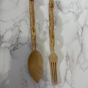 Könnte beinhalten: Ein hölzernes Löffel- und Gabel-Set. Beide Utensilien sind aus hellem Holz geschnitzt und weisen auf den Griffen komplizierte Designs auf. Der Löffel hat eine große, ovale Schüssel, während die Gabel vier Zinken hat. Das Set wird auf einer marmorierten Oberfläche präsentiert.
