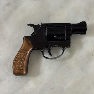 May include: A small toy revolver featuring a black metal frame and a textured bronze-coloured grip. The toy gun has a visible hammer, a trigger guard, and a rotating cylinder design, showing signs of paint wear along the edges of the frame.
