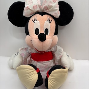 May include: A plush Minnie Mouse doll, featuring black ears, a pink nose, and a white and pink patterned bow. She wears a kimono-style outfit with a red sash and cream-colored shoes.