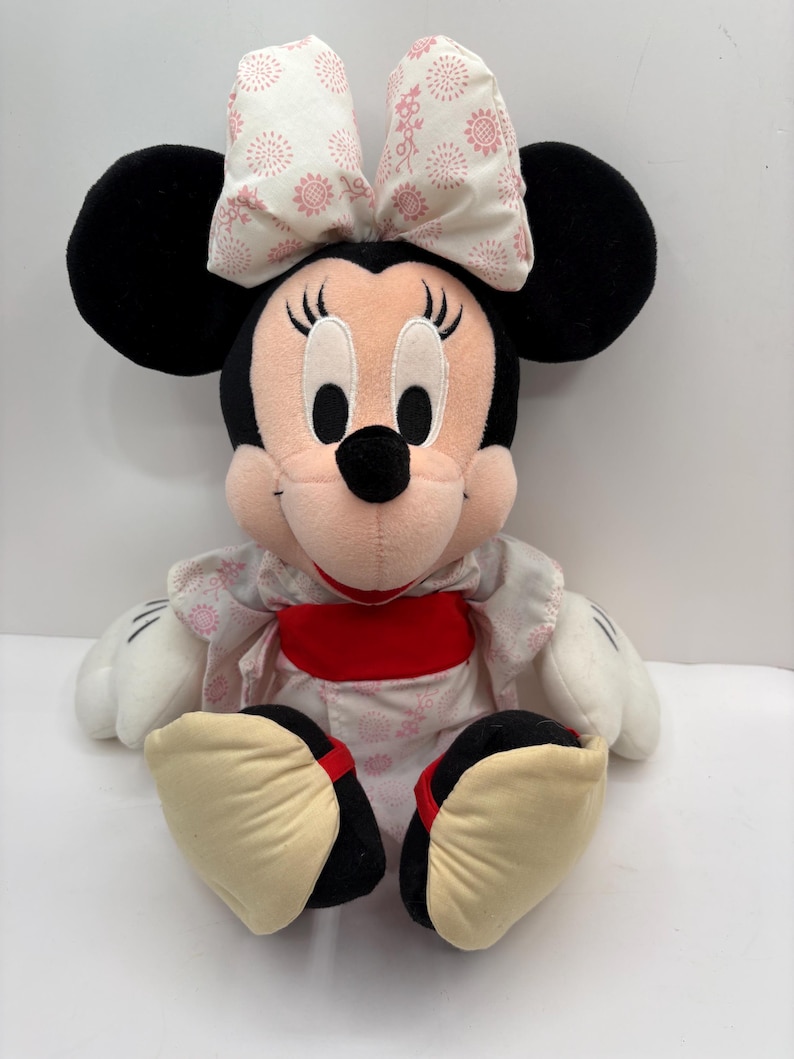 May include: A plush Minnie Mouse doll, featuring black ears, a pink nose, and a white and pink patterned bow. She wears a kimono-style outfit with a red sash and cream-colored shoes.