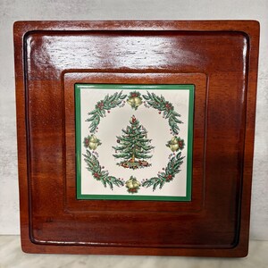 May include: A square, wooden serving tray with a recessed center. The center features a ceramic tile with a Christmas tree design surrounded by a wreath of greenery, bells, and berries. The tray is stained a rich, dark brown.