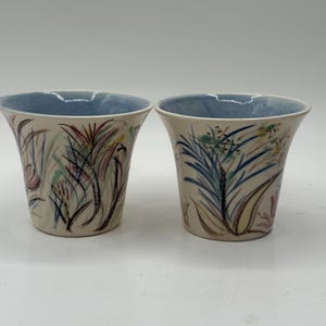 Puede incluir: Dos tazas de cerámica con interior azul claro y exterior beige. El exterior está decorado con un colorido diseño floral en tonos azules, verdes, rojos y amarillos. Las tazas tienen forma acampanada.