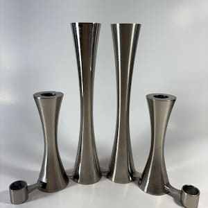 May include: A set of four brushed metal candle holders. Two tall, slender holders and two shorter, curved holders with attached side pieces. The metal has a silver-gray finish, reflecting light.
