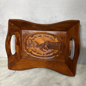 May include: A rectangular wooden tray with a carved central panel featuring a rural scene and the text "GIVE US THIS DAY OUR DAILY BREAD." The tray has curved sides, cut-out handles, and a warm brown finish.