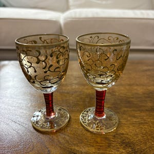 Könnte beinhalten: Zwei Vintage-Likörgläser mit bernsteinfarbenen Schalen und roten Stielen. Die Gläser haben goldene Filigranmuster und goldene Ränder. Die Böden sind ebenfalls mit goldenen Mustern verziert. Auf einer Holzoberfläche.