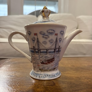 May include: A white ceramic teapot with a gold crown on the lid. The teapot features a blue and white illustration of the Albert Bridge in London, with the text "Thames Riverside London".
